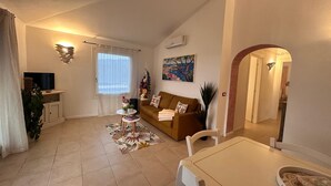 A living room with a sofa, a coffee table, a TV, and a dining area with a table and chairs. - EA WHITE VIBES (Baja Sardinia)