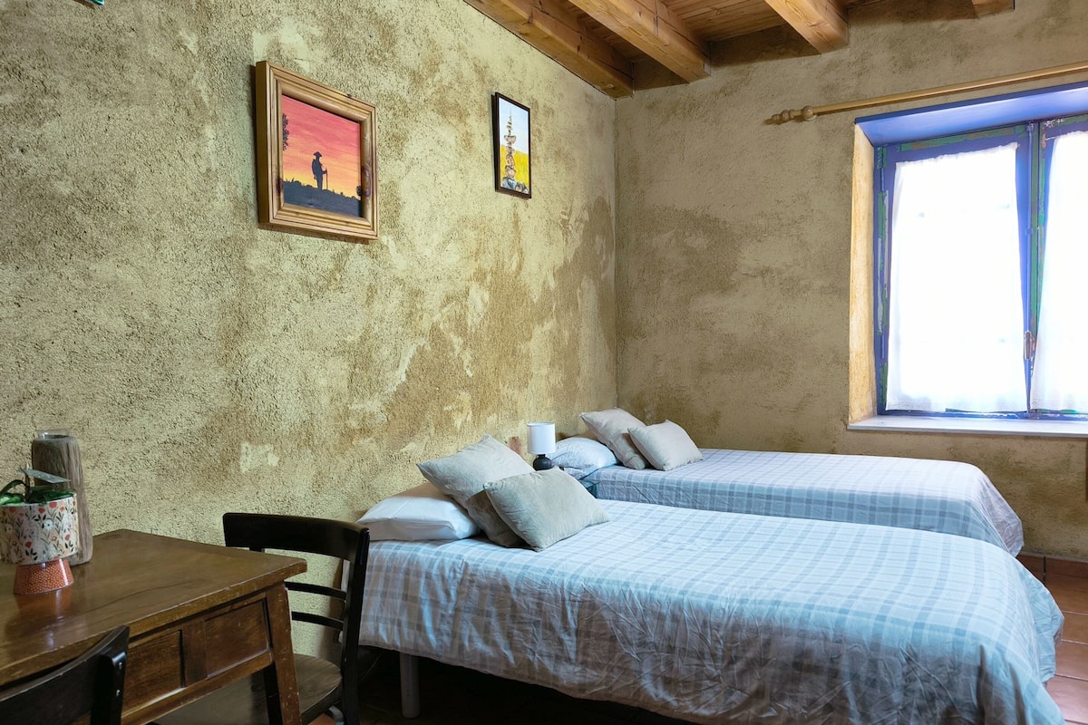 Una habitación con dos camas, una mesa de madera y una silla. Hay dos cuadros en la pared.
