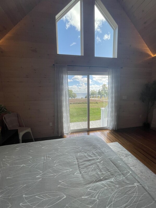 A bedroom with a large bed, a view of the outdoors through a sliding glass door, wooden walls, and a sloped ceiling.