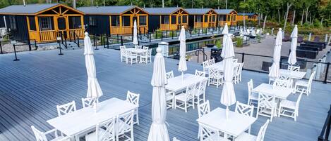 Una terraza con mesas y sillas blancas, una piscina y cabañas de madera con detalles amarillos.