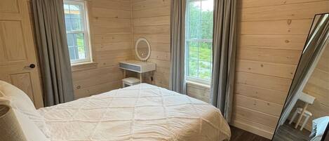 A wooden cabin bedroom with a bed, a bedside table, a mirror, and a window with curtains.