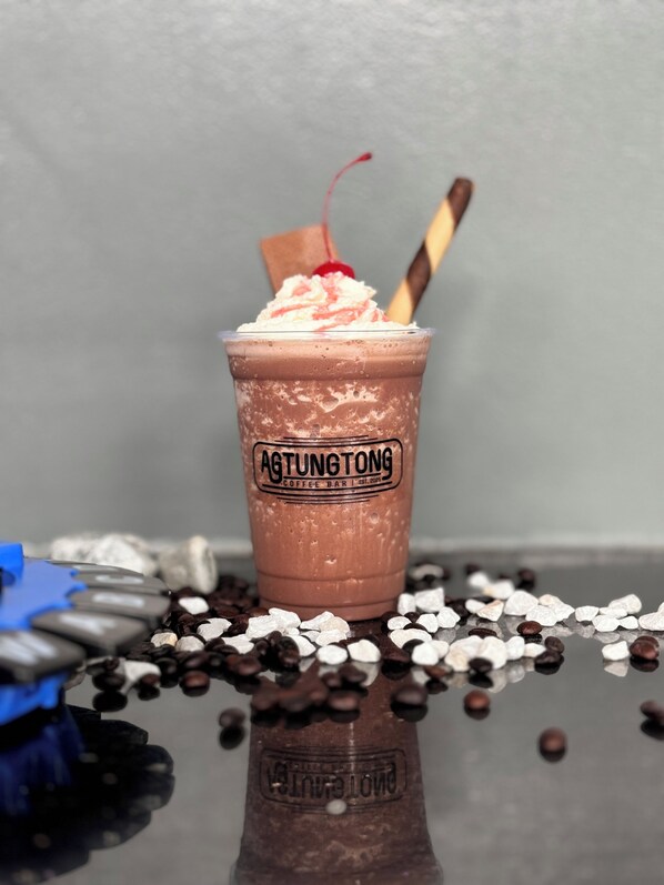 A cup of chocolate drink with whipped cream and a cherry on top, labeled "AGTUNG TONG COFFEE BAR EST. 2006".