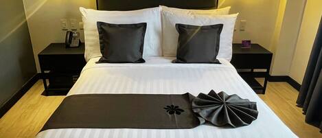 Una cama impecablemente tendida con sábanas blancas y dos almohadones oscuros, un cabecero negro y dos mesitas de noche con lámparas.