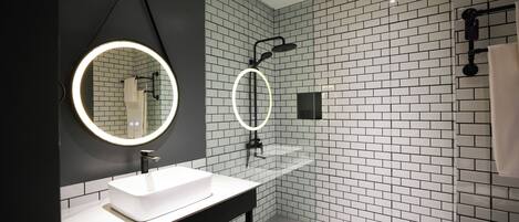 Une salle de bain moderne avec un lavabo blanc, un plan de travail noir et un miroir doté d’un éclairage circulaire. Les murs sont recouverts de carreaux blancs et noirs.