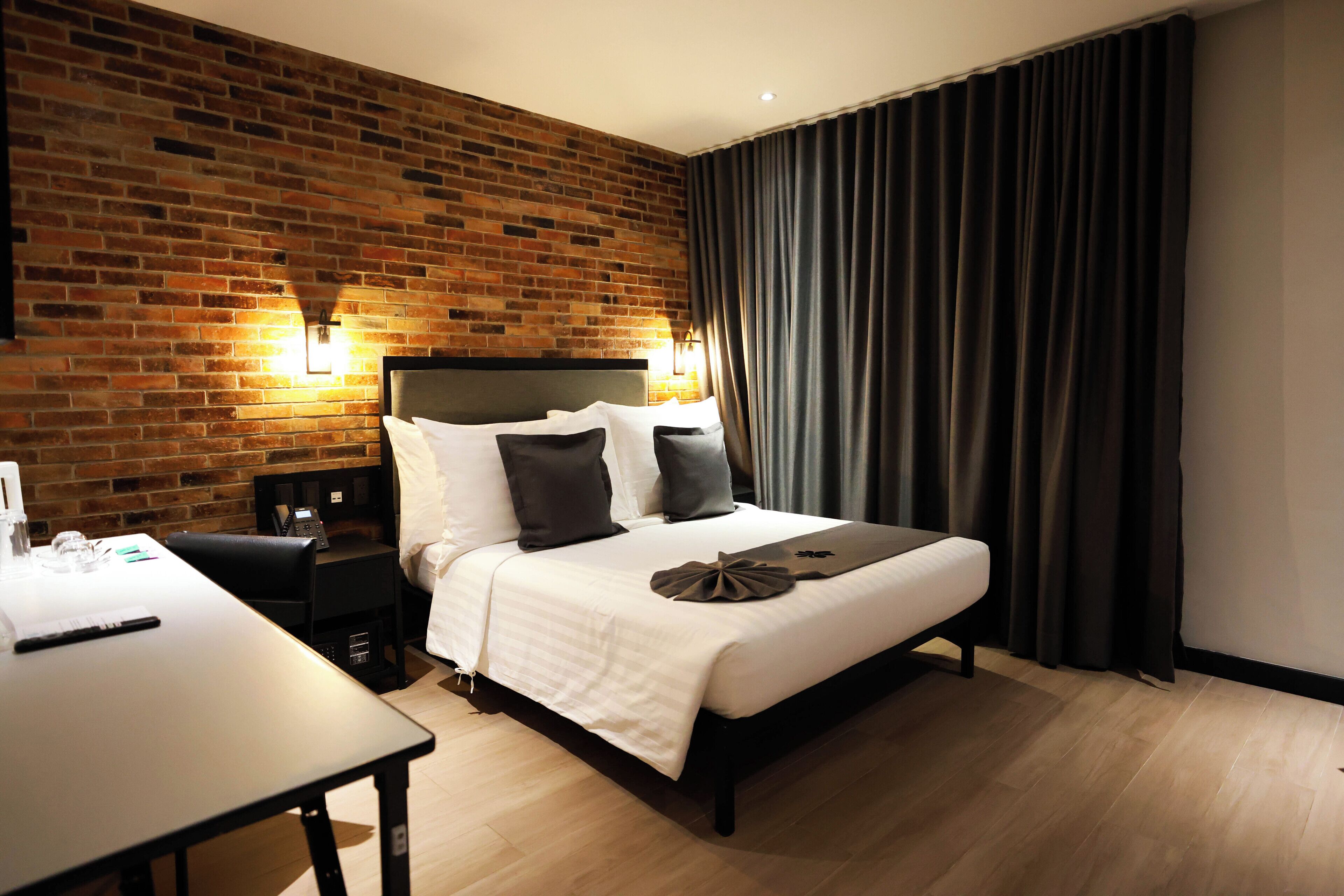A hotel room with a brick wall, a bed with white bedding and multiple pillows, a bedside table with a lamp, and a large window with dark curtains.