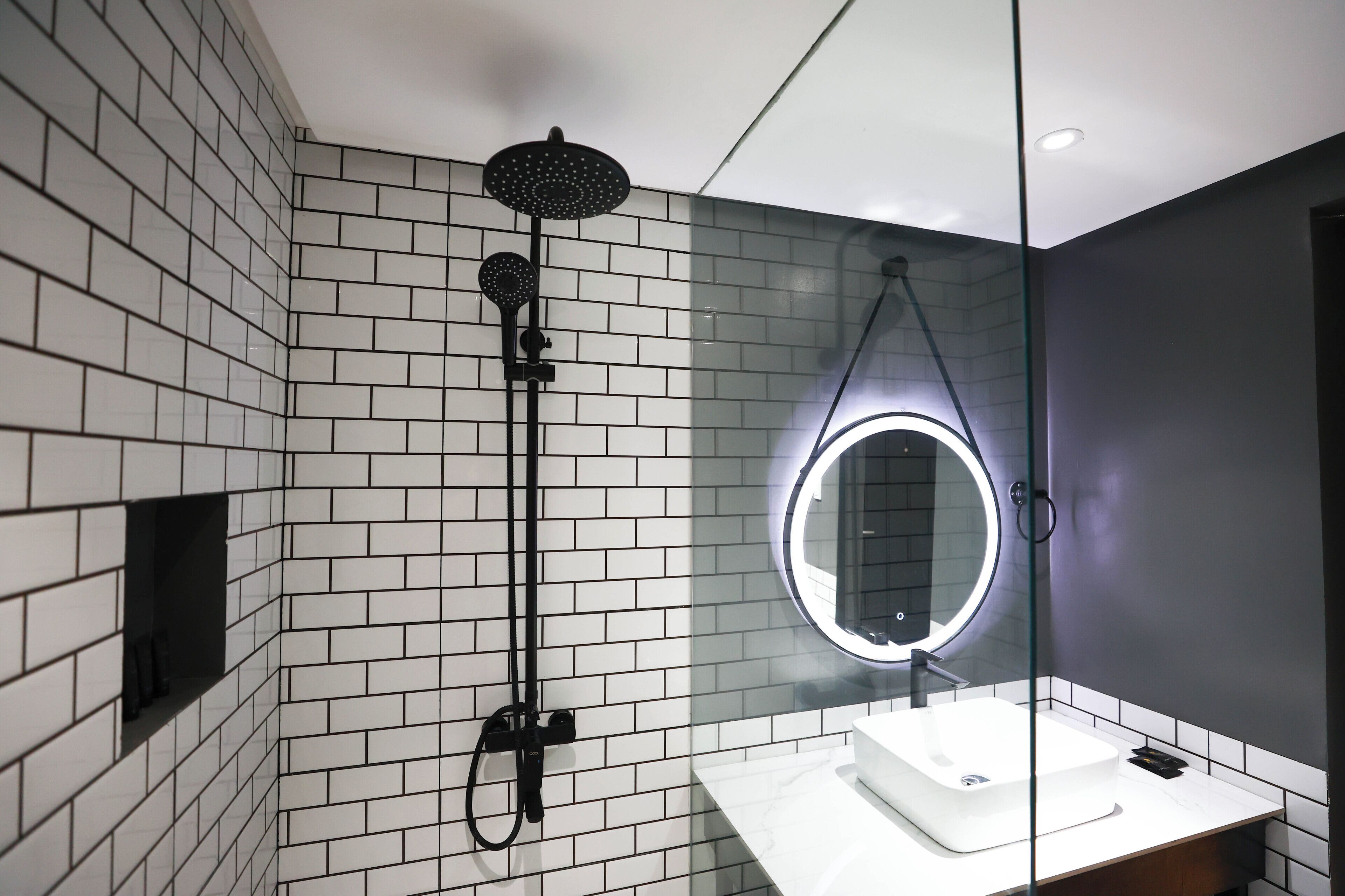 Un baño moderno con pared de azulejos, cabina de ducha de vidrio, espejo redondo con luz y lavamanos blanco.
