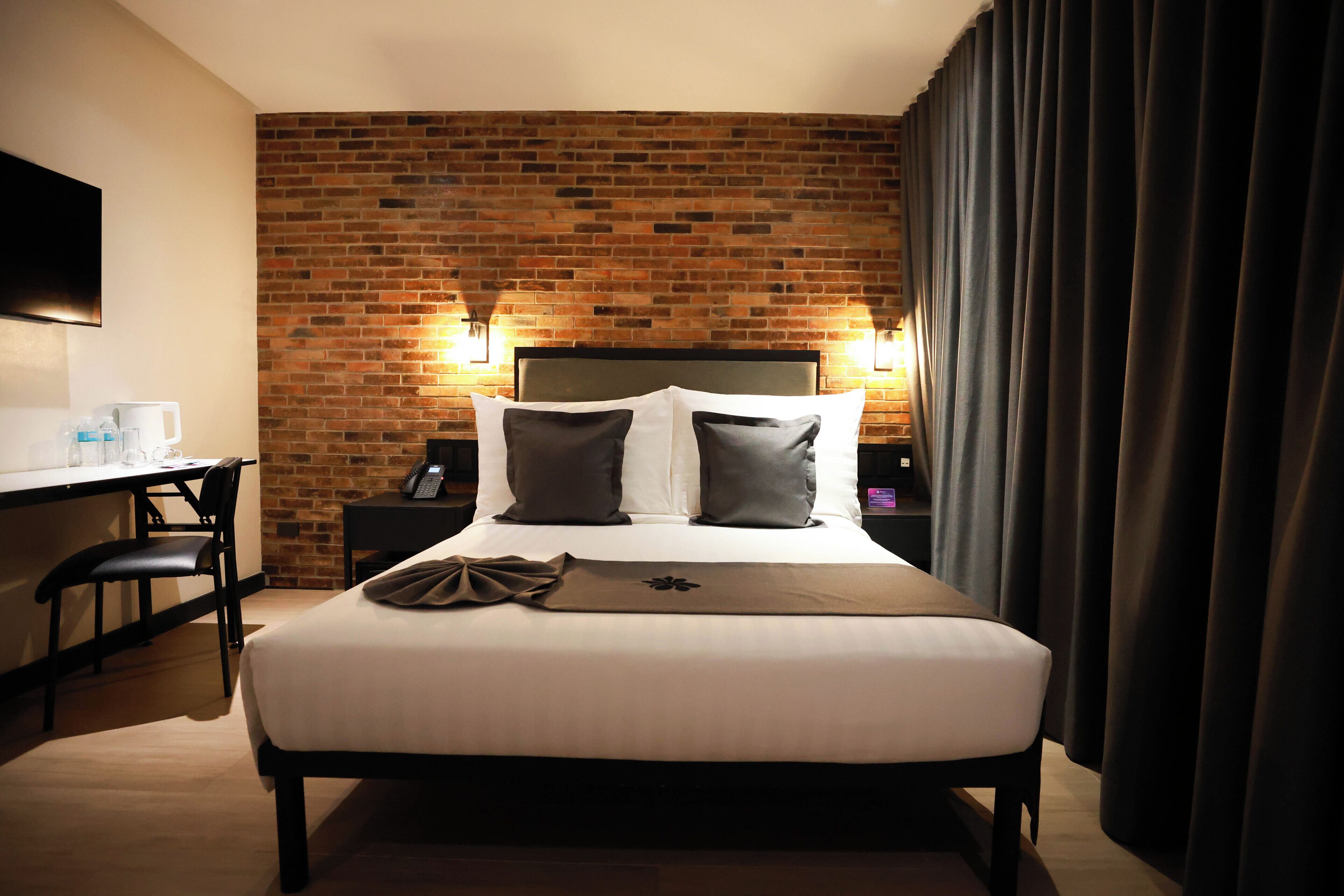 A hotel room with a brick wall, a bed with white bedding and two pillows, a desk with a chair, and a flat-screen TV mounted on the wall.