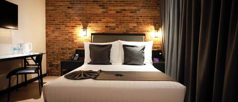 A hotel room with a brick wall, a bed with white bedding and two pillows, a desk with a chair, and a flat-screen TV mounted on the wall.