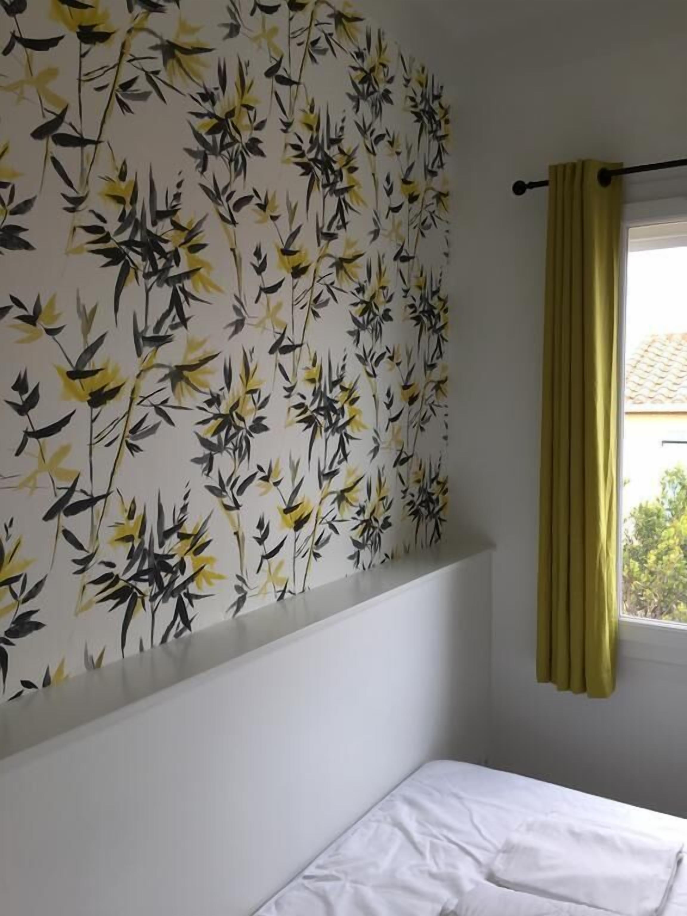 A room with a bed, a window with yellow curtains, and a wall with a floral pattern.