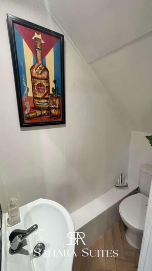 Une salle de bain avec un lavabo blanc, une œuvre d’art encadrée représentant une bouteille et des toilettes.