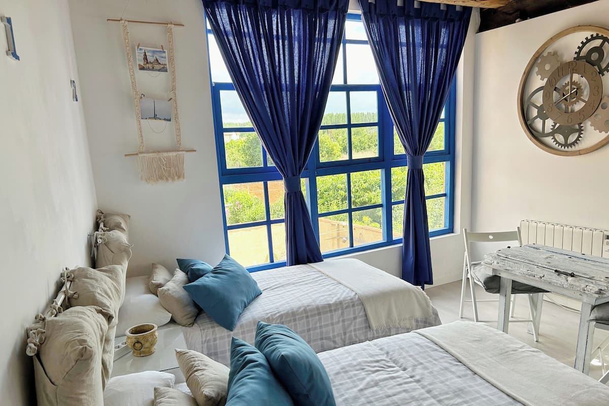 Un dormitorio con dos camas, un ventanal con cortinas azules, un reloj de pared y un radiador blanco.