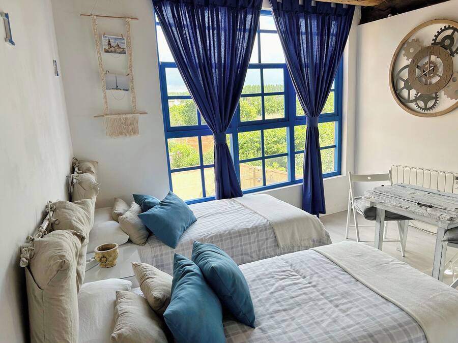 A bedroom with two beds, a large window with blue curtains, a wall clock, and a white radiator.