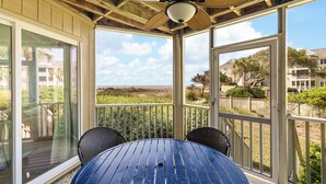 Ein überdachter Außenbereich mit einem runden Tisch, zwei Stühlen, einem Deckenventilator und großen Fenstern mit Blick auf den Strand und die umliegenden Gebäude.