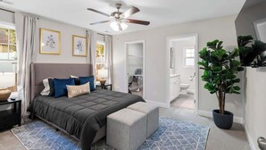 Une chambre à coucher avec un lit, un ventilateur de plafond, une plante en pot et une salle de bain visible à travers une porte ouverte.