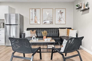 Uma sala de estar moderna com um sofá preto, duas cadeiras pretas, uma mesa de centro com objetos decorativos, uma geladeira e arte na parede retratando cenas tropicais.