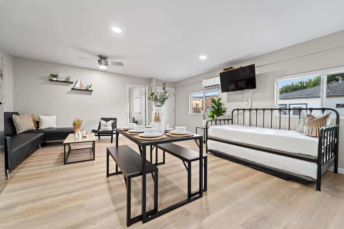 Una sala moderna con un sofá negro, un sofá blanco, una mesa de comedor con individuales, un banco y una cama con estructura de metal.