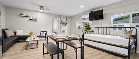 Una sala moderna con un sofá negro, un sofá blanco, una mesa de comedor con individuales, un banco y una cama con estructura de metal.