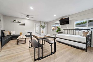 A modern living room with a black sofa, a white couch, a dining table with placemats, a bench, and a bed with a metal frame.