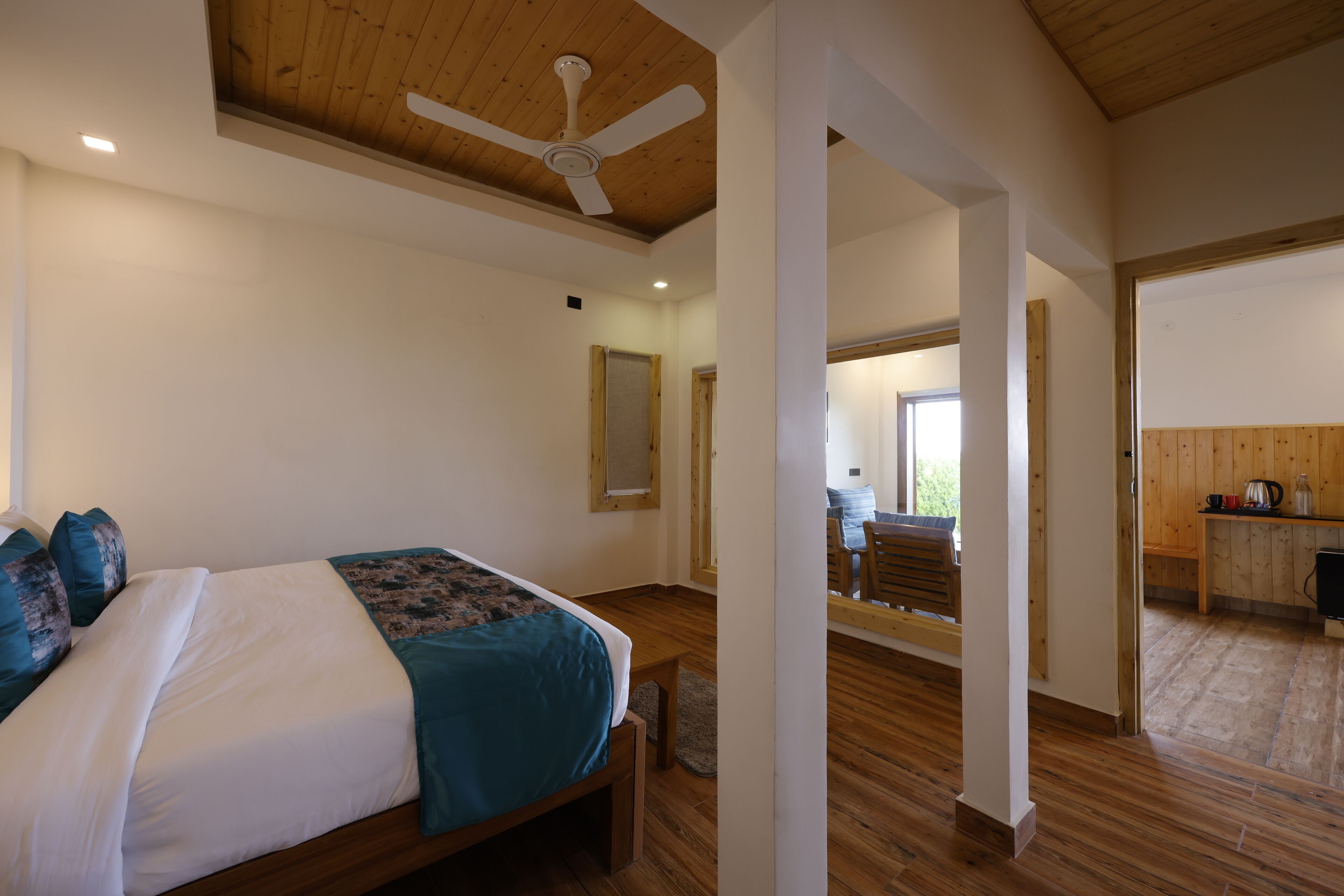 A bedroom with a bed, wooden floor, ceiling fan, and a view into the dining area.