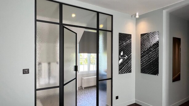 A modern interior with a glass door leading to a bright room, a radiator, and two abstract art pieces on the wall. - Le Petit Château (Baye)