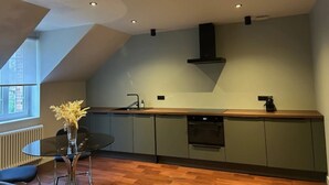 A modern kitchen with a built-in oven, a dishwasher, and a black range hood. There is a round dining table with a vase of flowers and a chair. The kitchen has a wooden countertop and a window providing natural light. - Le Petit Château (Baye)