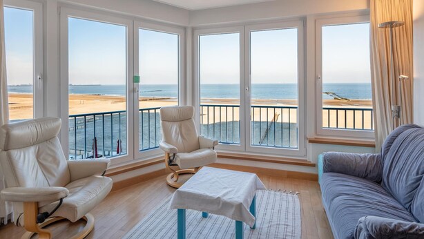 Ein geräumiges Wohnzimmer mit großen Fenstern und Blick auf einen Strand, ausgestattet mit einer Couch, Sesseln und einem kleinen Tisch.
