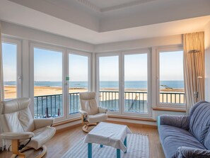 Ein geräumiges Wohnzimmer mit großen Fenstern und Blick auf einen Strand, ausgestattet mit einer Couch, Sesseln und einem kleinen Tisch.