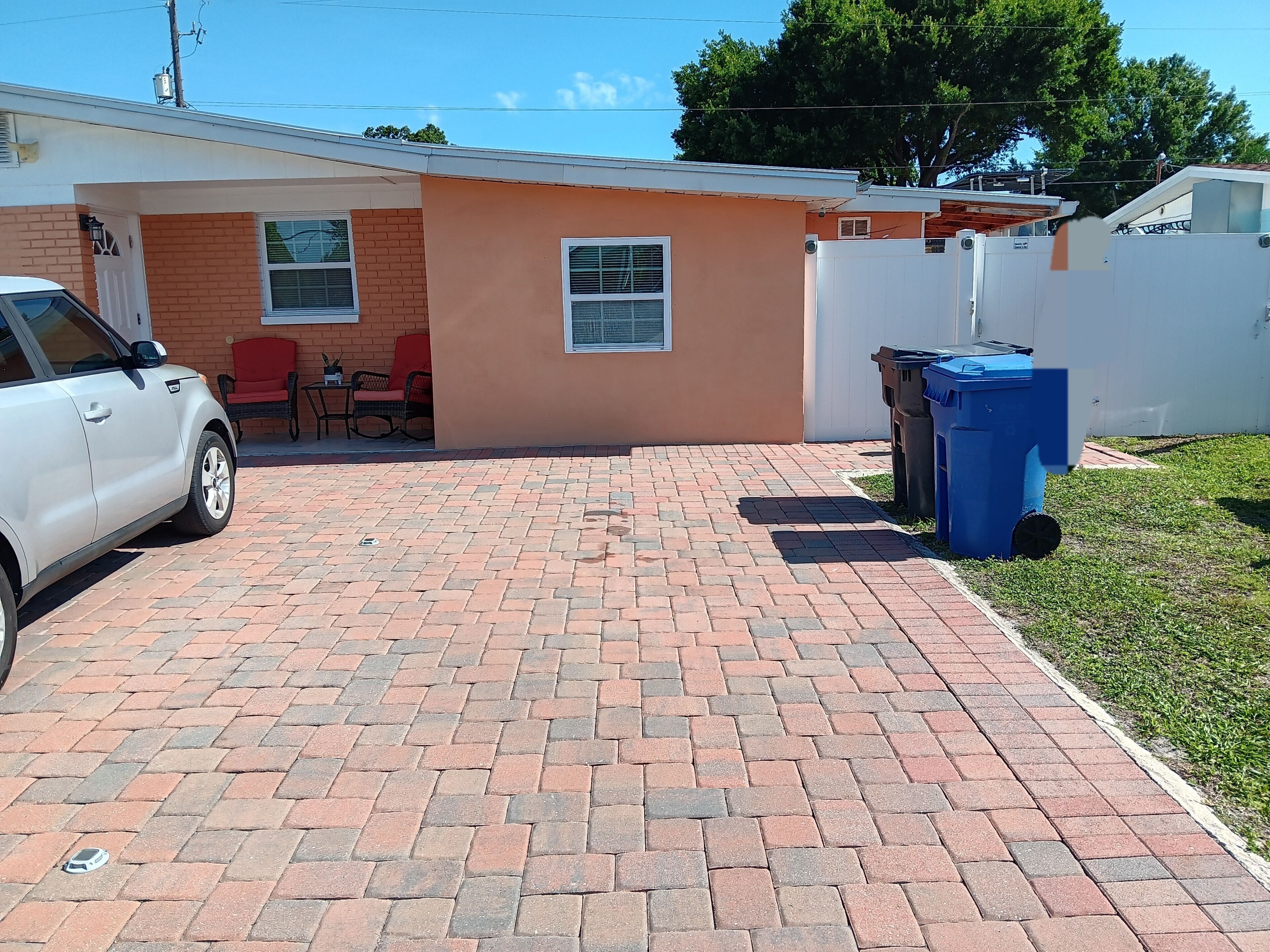 A residential driveway with a white car parked on the left, a brick patio, a small red chair, and a white fence.
