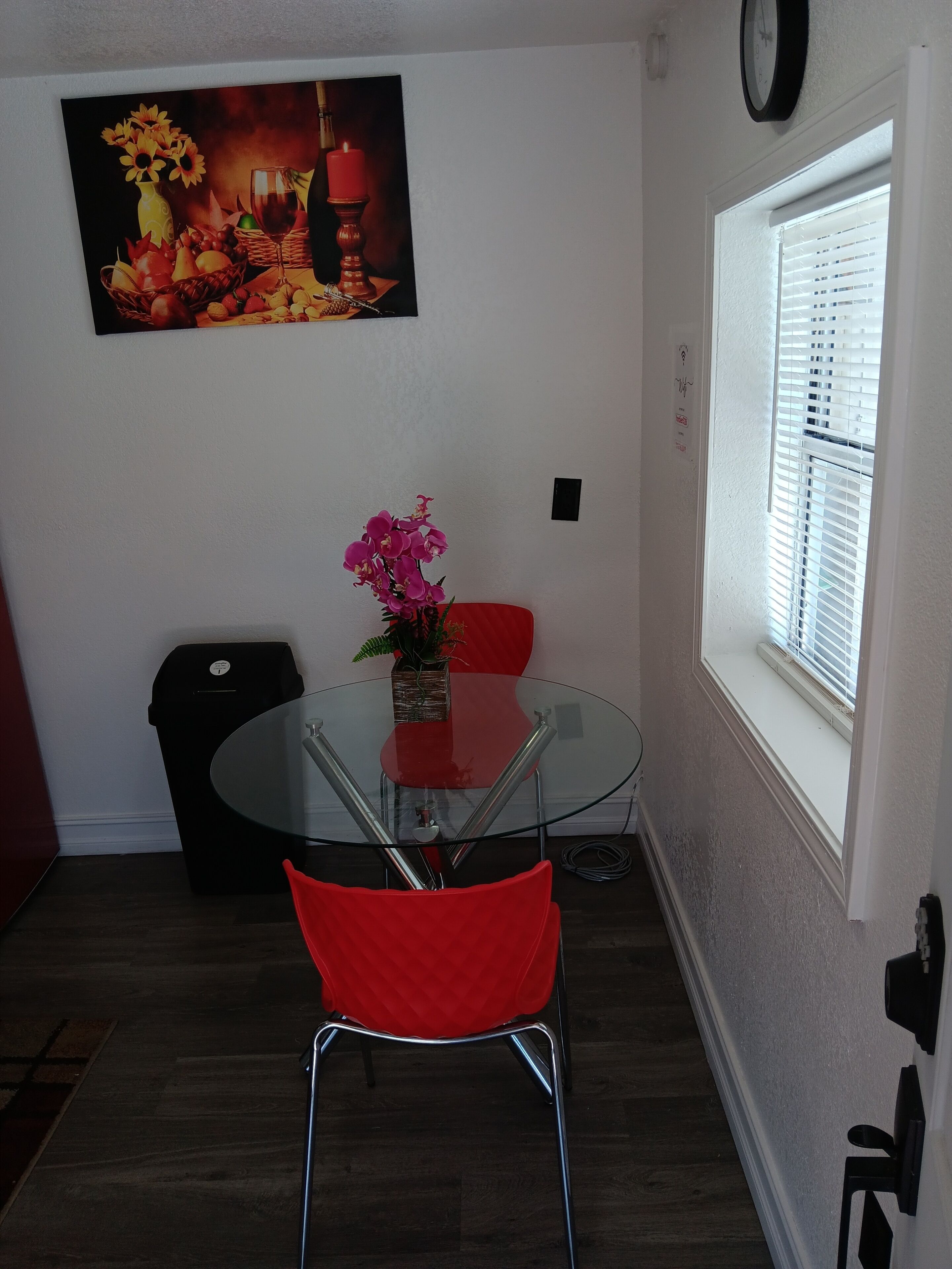A dining area with a glass table, red chairs, a vase with flowers, a painting on the wall, and a window with blinds.