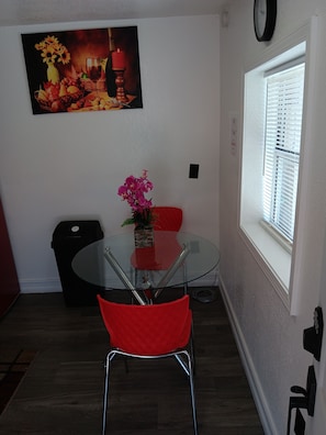 Un coin repas avec une table en verre, des chaises rouges, un vase de fleurs, un tableau au mur et une fenêtre avec des stores.
