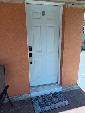 A white door with a number plate, a doormat with a welcome message, and a chair outside.