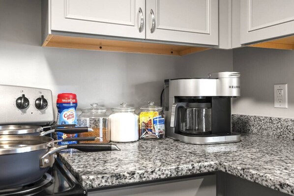 A kitchen with a stove, coffee maker, and various food items on the counter.