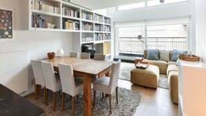 A modern living room with a wooden dining table, white chairs, a beige sofa, and a large bookshelf filled with books. - Duplex at Albert Beach With Stunning Sea Views (Knokke-Heist)