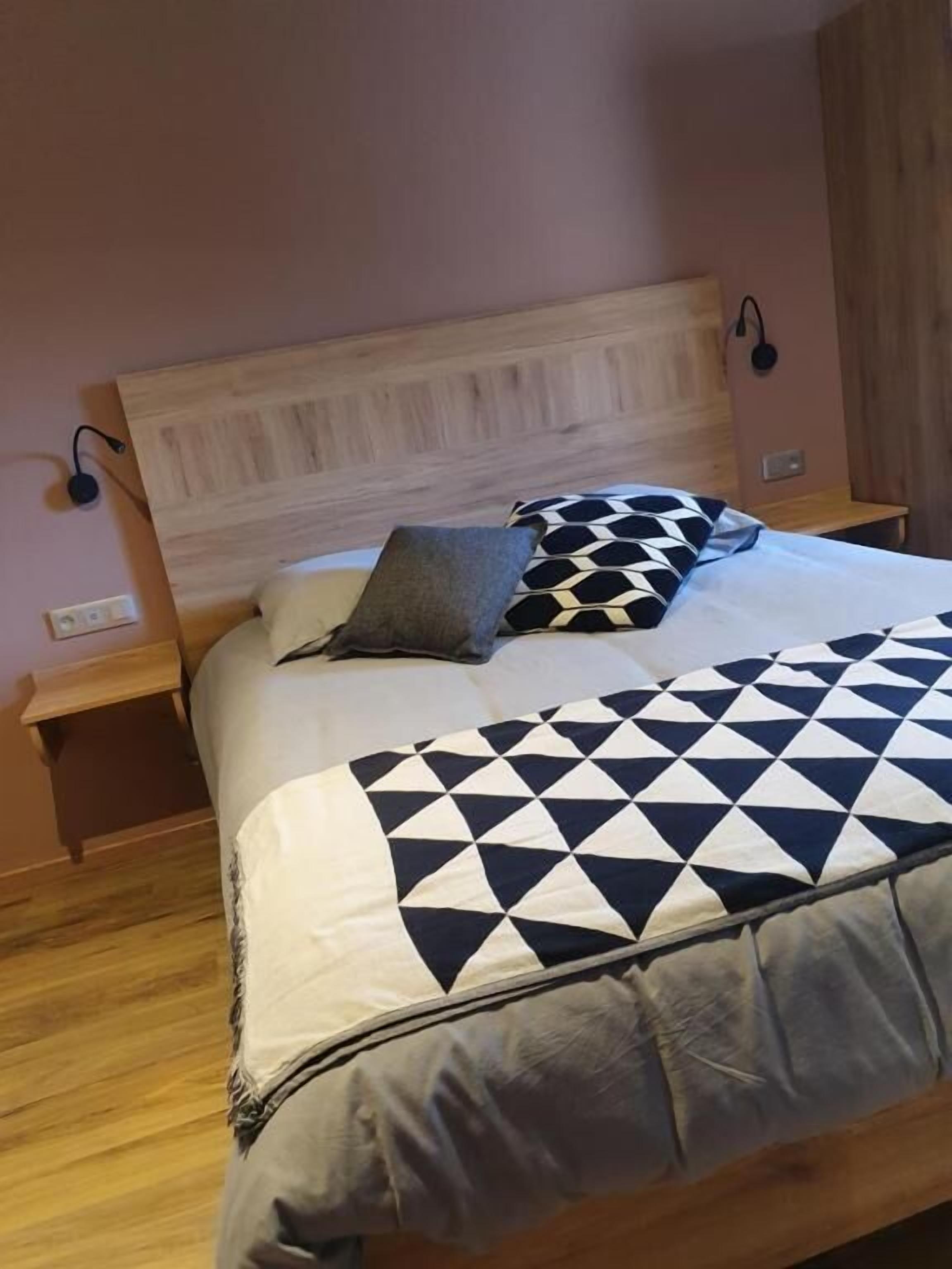 A bedroom with a wooden headboard, a bed with a geometric patterned bedspread, two bedside tables, and a wall-mounted lamp.
