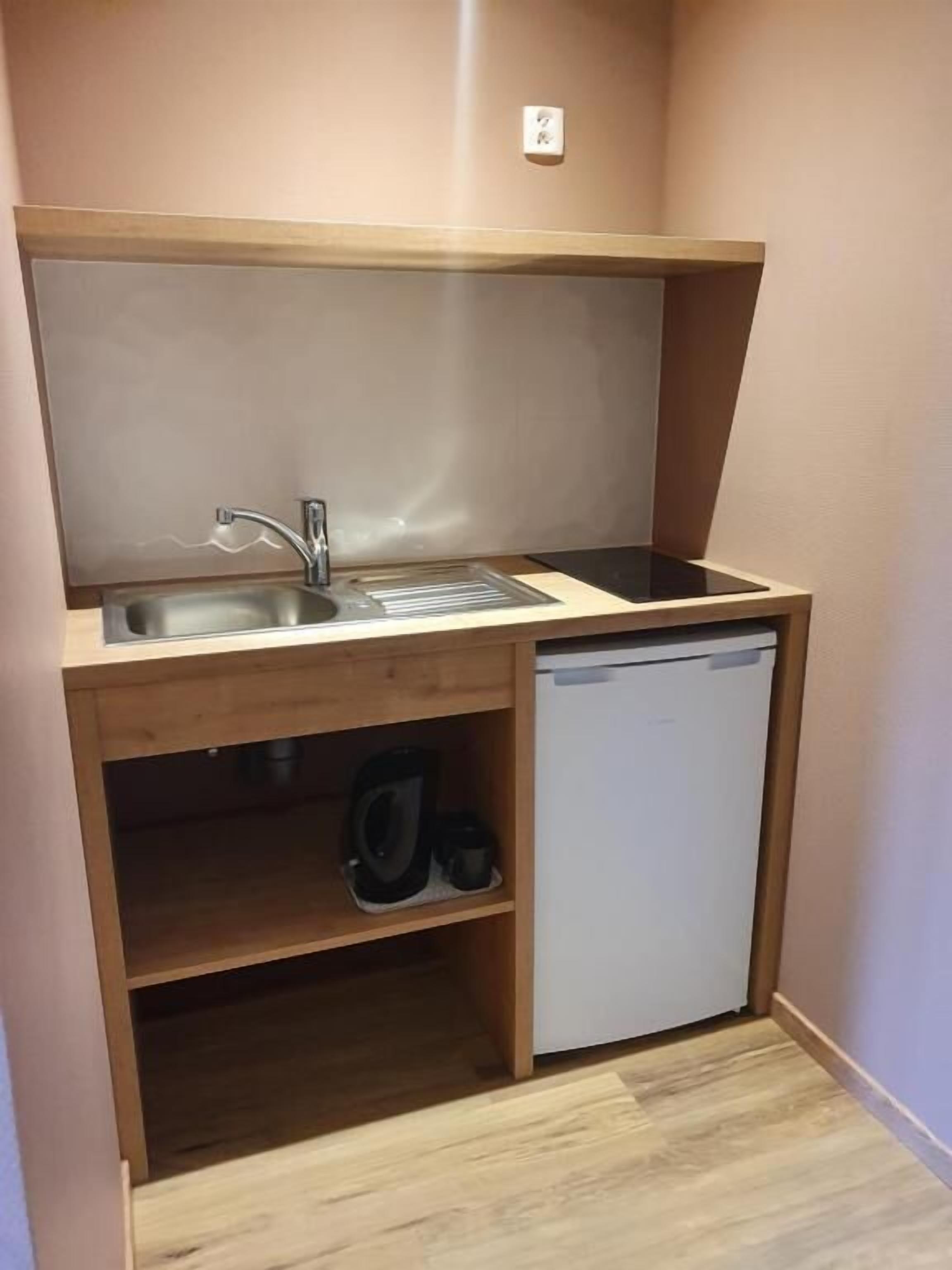 Una kitchenette compacta con un lavamanos, un refrigerador pequeño y una alacena con una tetera y tazas.