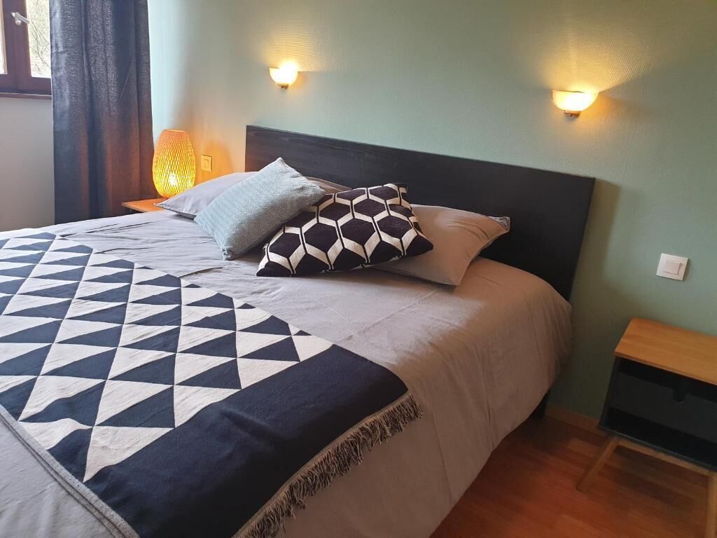 Una cama bien hecha con un edredón estampado con motivos geométricos y varias almohadas, una mesita de noche con una lámpara y un aplique de pared.