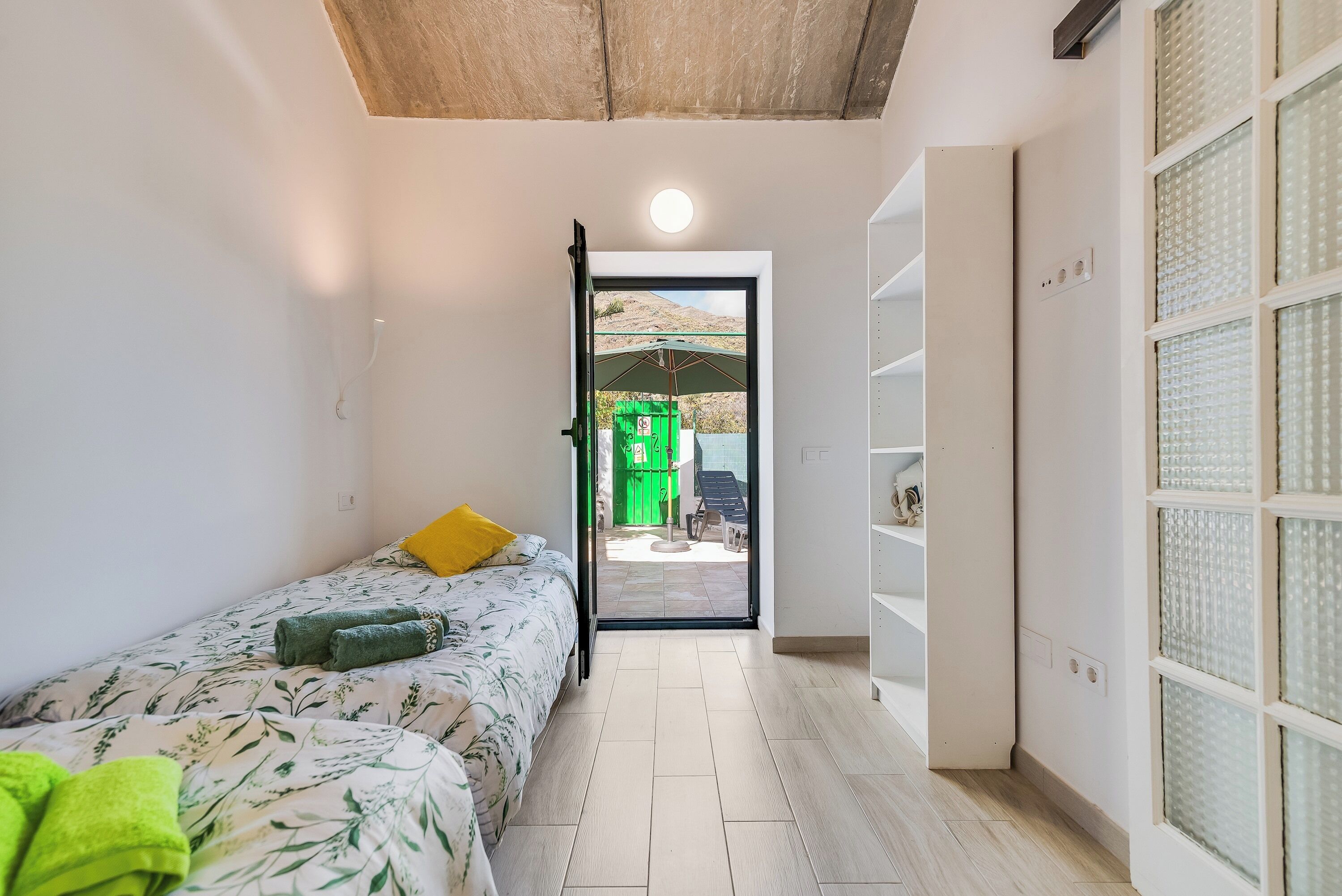 Un pasillo estrecho con una cama a la izquierda, una puerta que da a un espacio exterior y un estante blanco a la derecha.