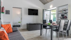 Ein modernes Wohnzimmer mit einer Couch, einem Esstisch und Stühlen. Ein Fernseher ist an der Wand befestigt, und es gibt eine Küchenzeile mit einem Kühlschrank und Regalen.