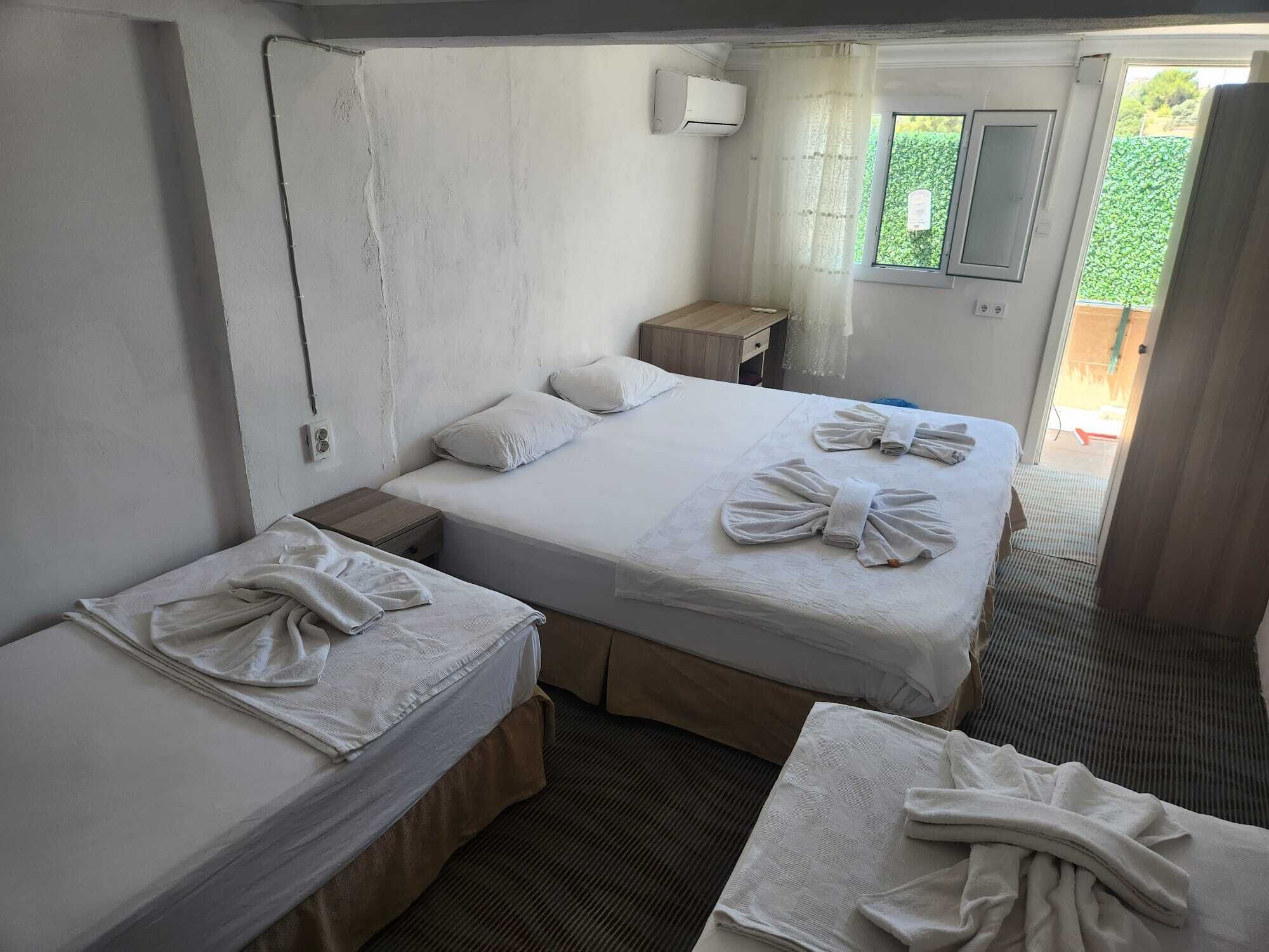 Une chambre d’hôtel avec deux lits, une petite table de chevet en bois, une fenêtre donnant sur un espace verdoyant et un climatiseur fixé au mur.