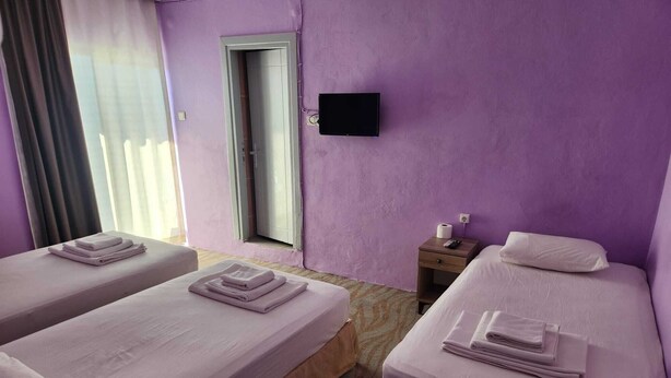 A hotel room with two beds, a TV on the wall, a window with curtains, and a small bedside table with a remote control. - Cesme MASAL (Cesme)