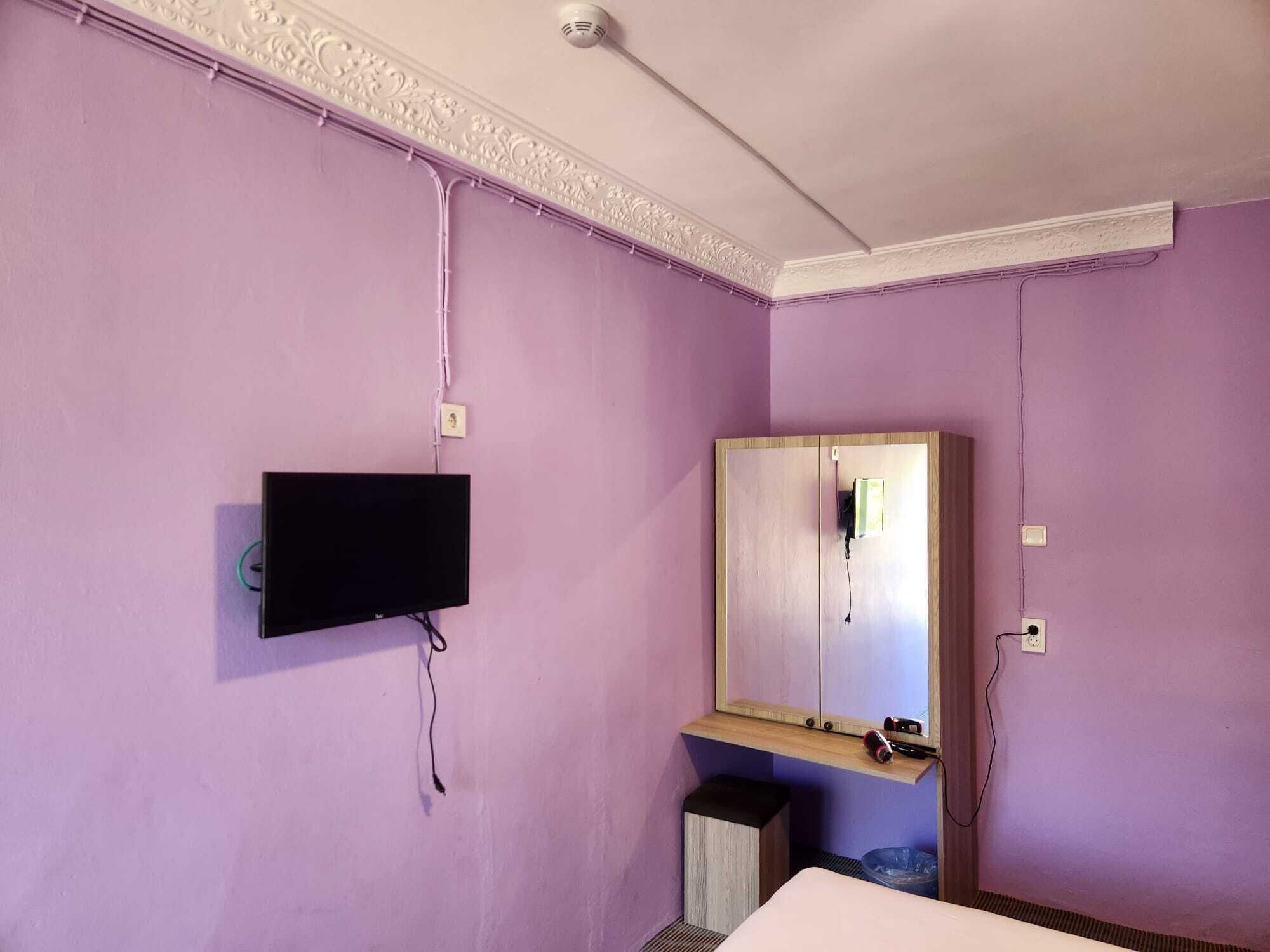Una habitación con paredes moradas, un televisor montado, un mueble de madera con espejo y un pequeño cubo de basura.
