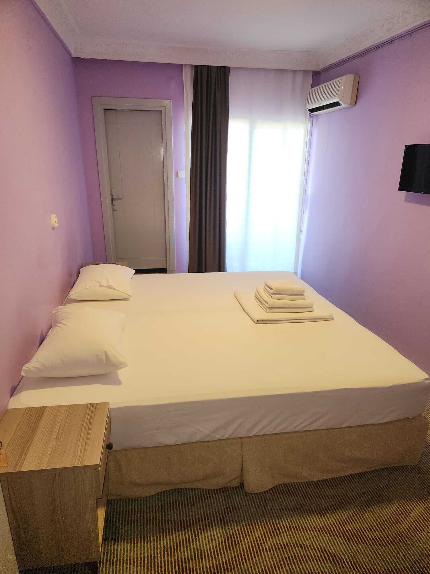 A hotel room with a single bed, a wooden nightstand, a wall-mounted TV, and an air conditioning unit.