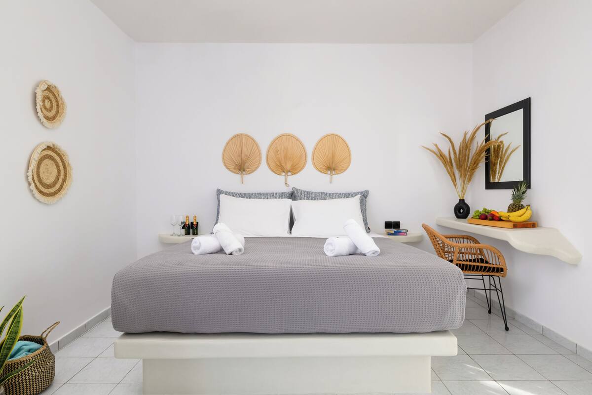 Quarto de hotel moderno com uma cama grande, uma mesa de cabeceira com uma garrafa e copos, uma cadeira e uma pequena prateleira com frutas e uma planta.