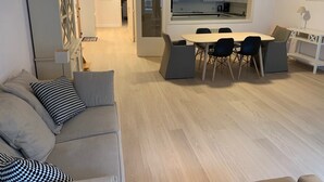 En moderne stue med sofa, spisebord med stole, og et køkken, der ses igennem et vindue.