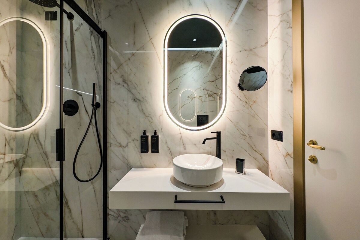 Un baño moderno con ducha de vidrio, lavamanos de mármol y dos espejos iluminados.