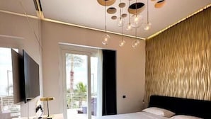 Ein modernes Schlafzimmer mit einem großen Bett, einem Schreibtisch und einem Sessel. An der Wand ist ein Fernseher angebracht und das Fenster bietet einen Blick ins Freie.