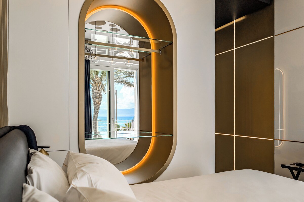 A modern hotel room with a curved mirror, a bed with white linens, and a view of the ocean through large windows.
