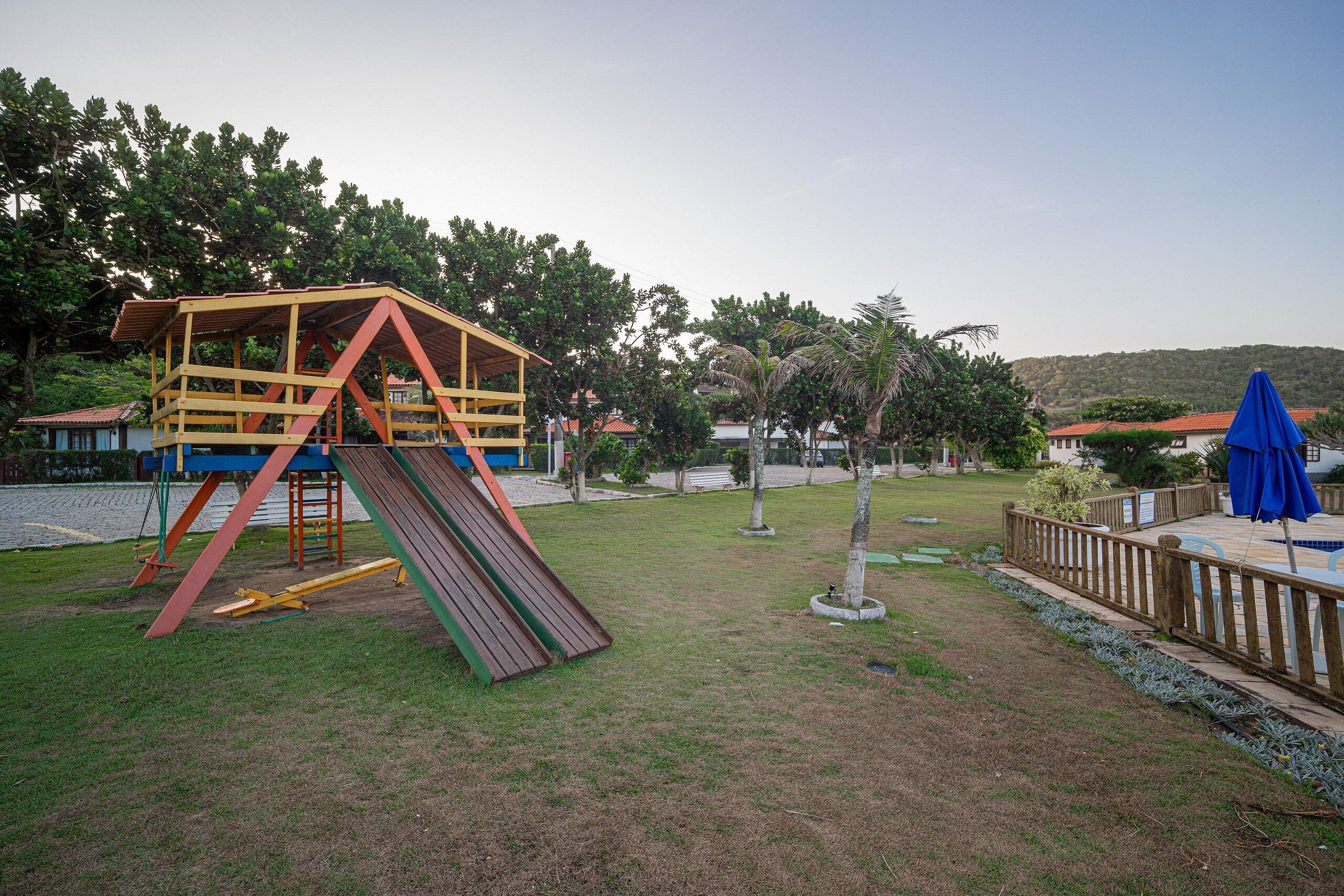 Ein Spielplatz mit Rutsche und Klettergerüst, umgeben von einem eingezäunten Bereich mit Bäumen und Blick auf Häuser im Hintergrund.
