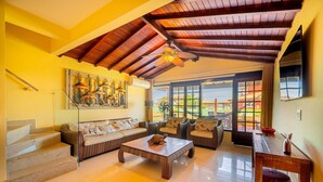 A spacious living area with a wooden ceiling, a staircase, a large painting, and a view through the glass doors. - Bz60 Casa em condomínio pé na areia (Buzios)