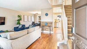Ein geräumiges Wohnzimmer mit einem großen beigefarbenen Sofa, einem blauen Sessel und einem Essbereich mit einem Holztisch und Stühlen. Eine Treppe führt zu einer oberen Ebene mit einem weißen Geländer.